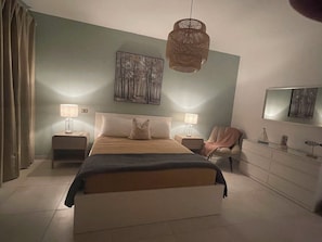 Un dormitorio con una cama, dos mesitas de noche, una silla, un aparador y una lámpara colgante.