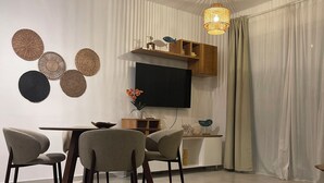 Ein moderner Essbereich mit einem runden Tisch, vier Stühlen, einem Flachbildfernseher, Wanddekoration und einer Pendelleuchte.