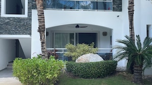 Ein modernes, zweistöckiges Gebäude mit Natursteinfassade, einem Balkon und einem gepflegten Garten mit Palmen und Sträuchern.