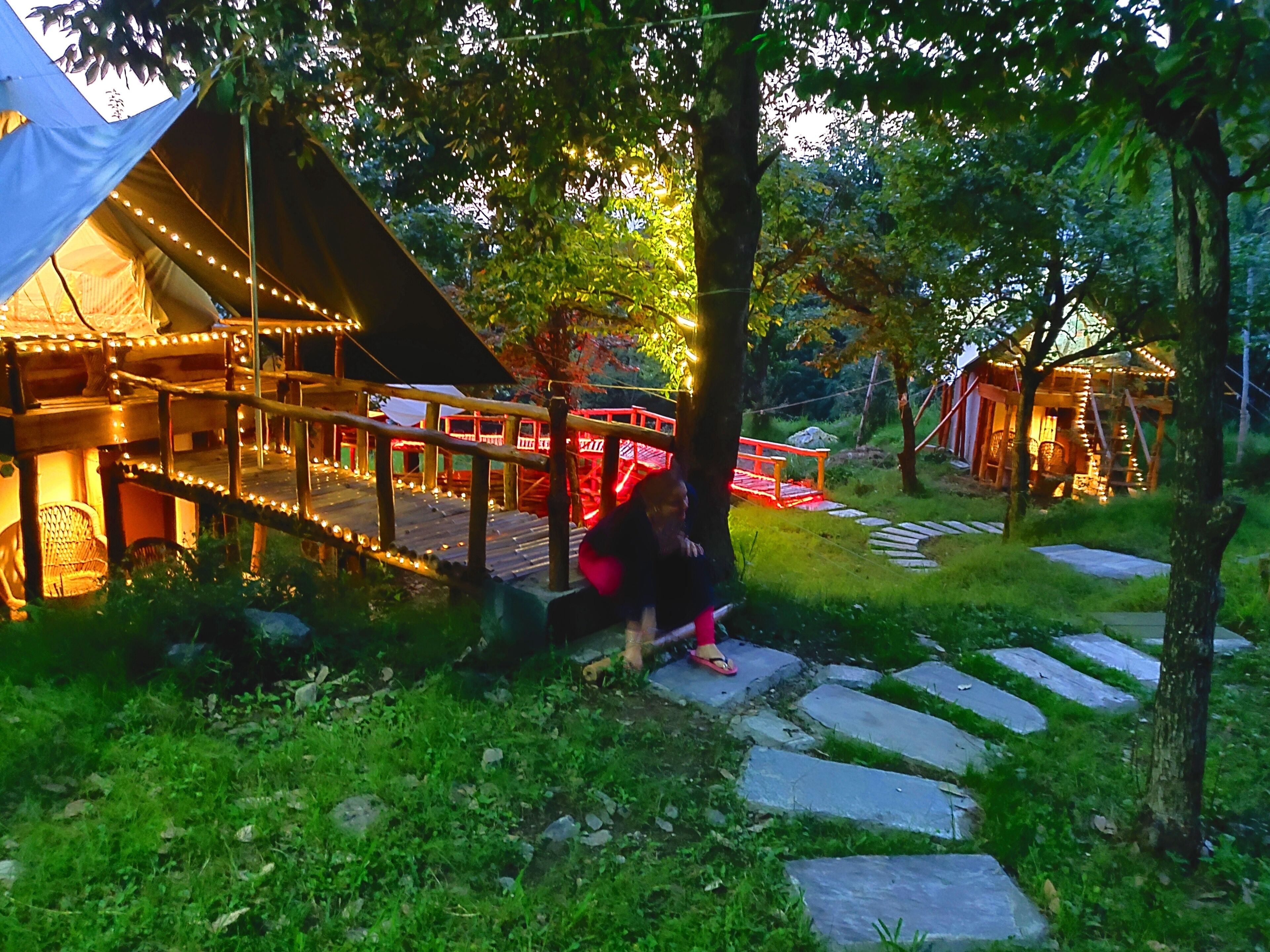 A tented area with a wooden deck and string lights, surrounded by greenery and stone pathways.