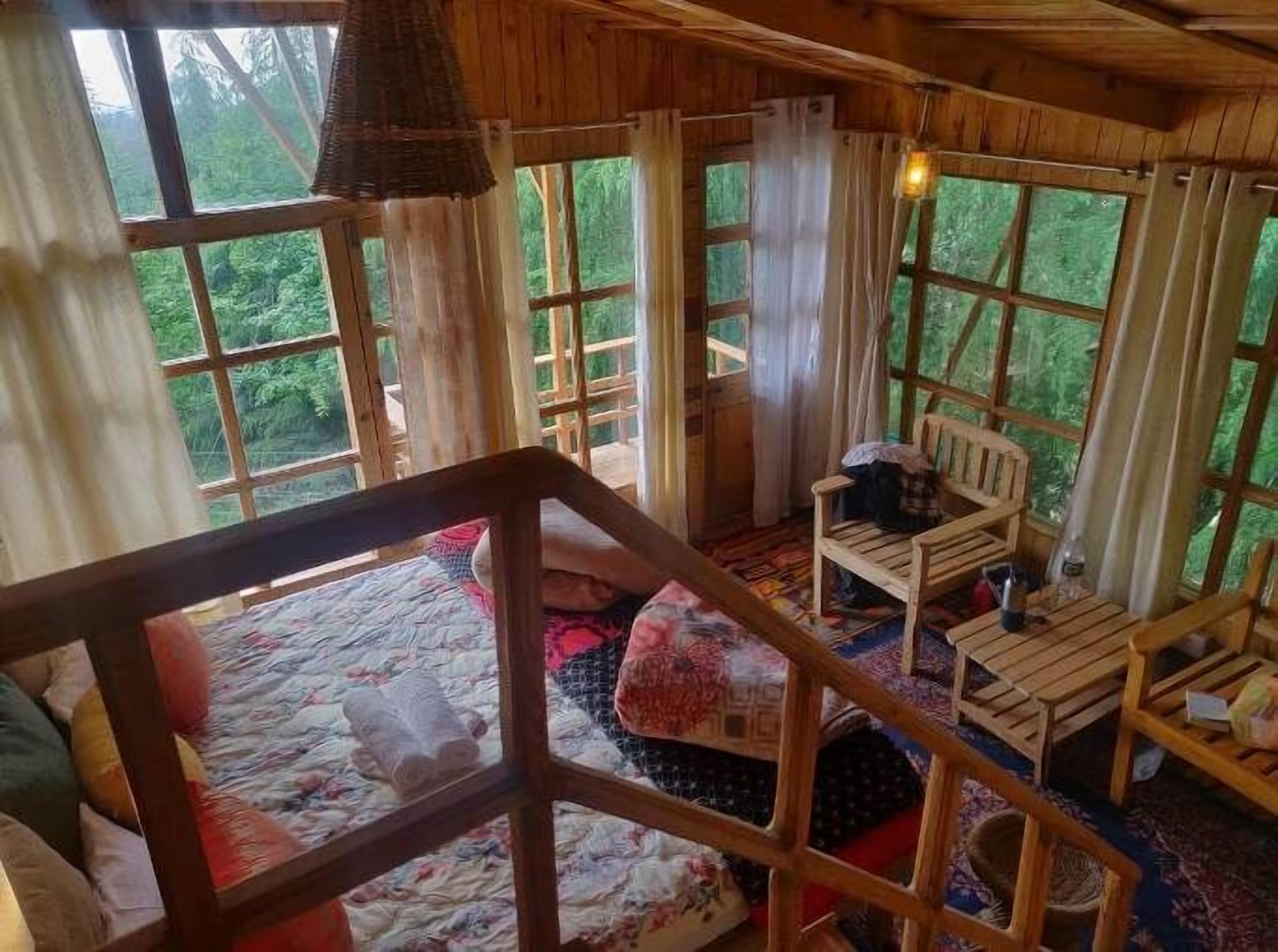 A cozy cabin interior with a wooden staircase, a bed with colorful bedding, a wooden table, and large windows offering a view of the forest.