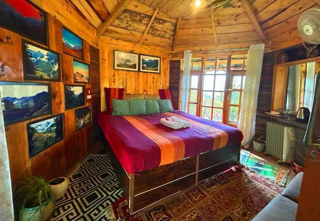 A wooden cabin bedroom with a bed, framed pictures on the walls, and a window with curtains.