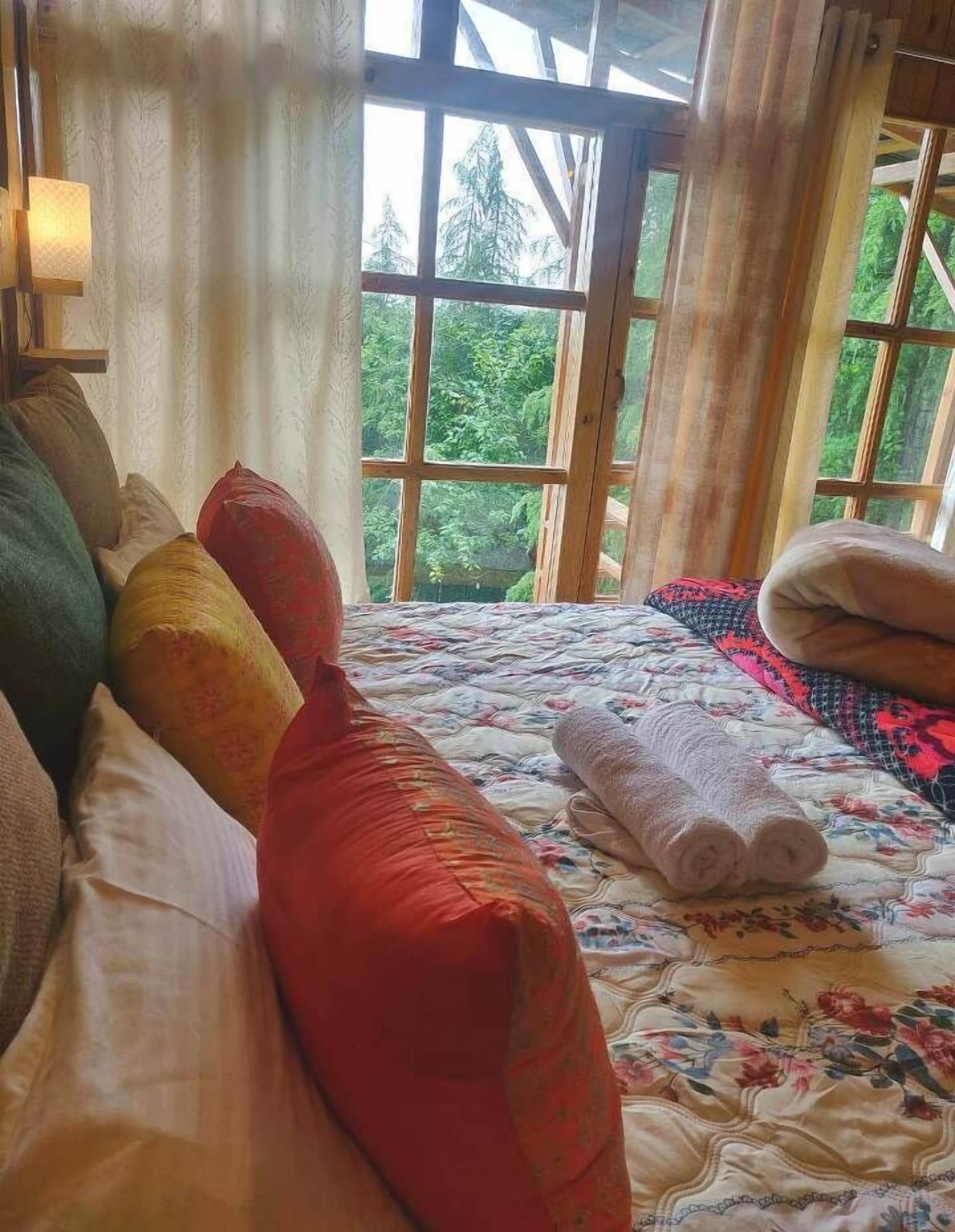 A bed with multiple pillows and a rolled towel, a window with a view of greenery, and a curtain.