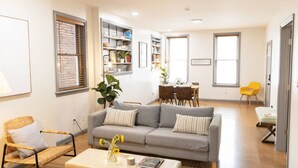 A modern living room with a grey sofa, a wicker chair, and a dining area with a table and chairs. - Chic Brownstone in Historic Troy w/Furnished Deck (Troy)