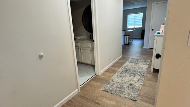 Een gang met houten vloer, een grijs vloerkleed en een witte deur die leidt naar een badkamer met een ronde spiegel.