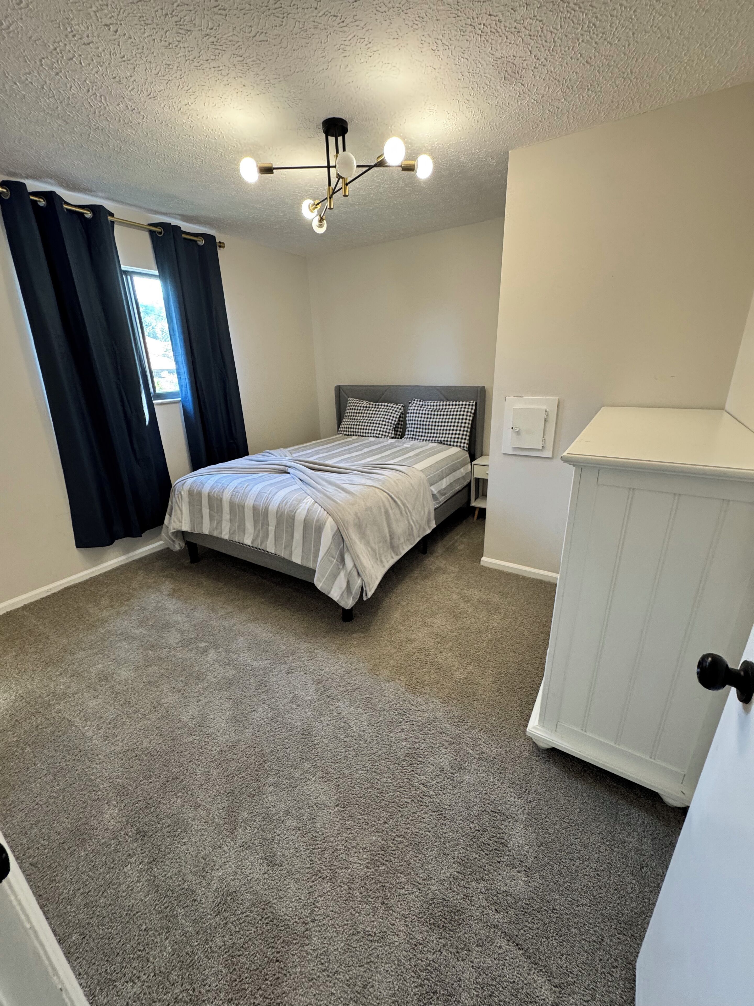 A bedroom with a bed, a nightstand, a dresser, a ceiling light fixture, and a window with curtains.
