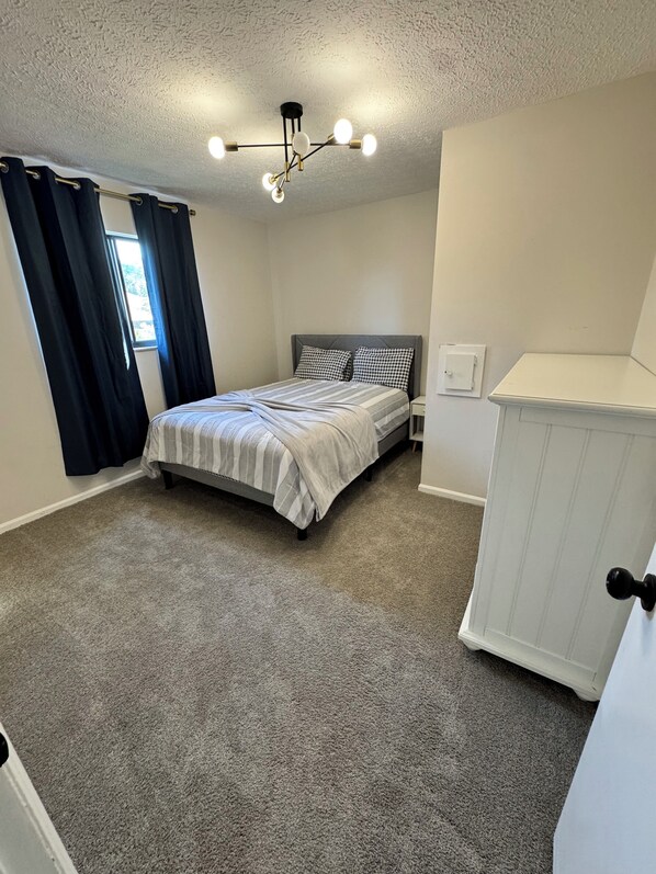 Een slaapkamer met een bed, een nachtkastje, een dressoir, een plafondlamp en een raam met gordijnen.