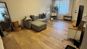 Ein geräumiges Wohnzimmer mit einem grauen Ecksofa, Holzboden, einem großen Spiegel und einem Fernseher auf einem Holztisch.