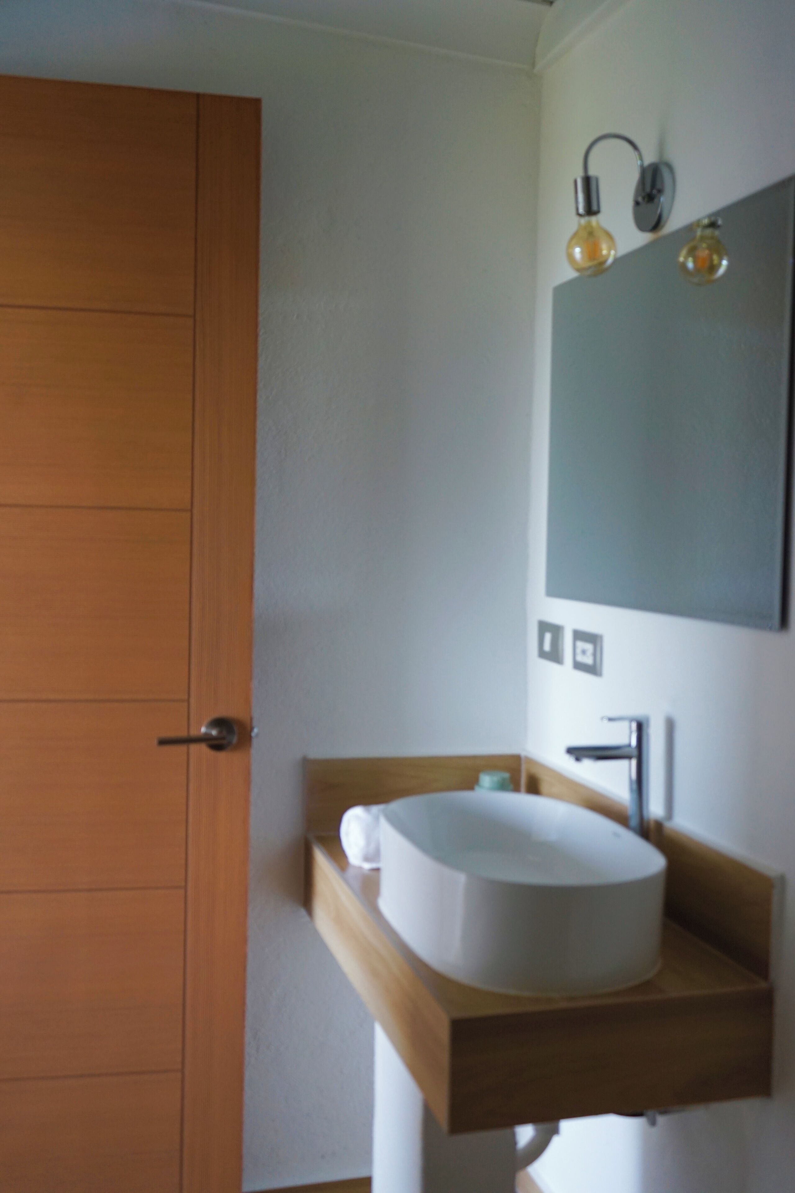 A bathroom with a wooden door, a white sink with a wooden countertop, and a mirror with two light fixtures.