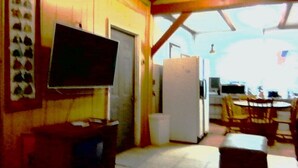 Una habitación con televisión montada en la pared, un refrigerador, una mesa de comedor con sillas y una ventana que deja entrar luz natural.