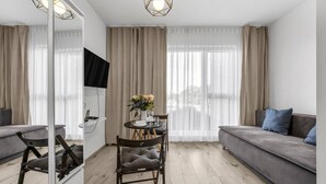 Een moderne woonkamer met een bank, een eettafel met stoelen, een aan de muur bevestigde televisie en grote ramen met gordijnen.