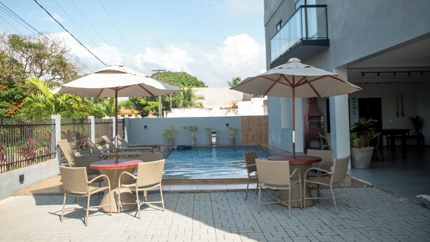 An outdoor pool area with wicker furniture and umbrellas, surrounded by a brick patio and a building with a balcony. - Apto c Piscina e 2 su tes a Poucos Passos da Praia (Japaratinga)