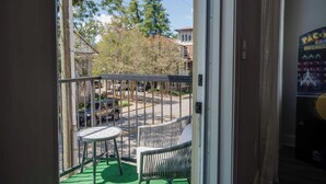 A balcony with a wicker chair and a small table, overlooking a street with parked cars and trees.