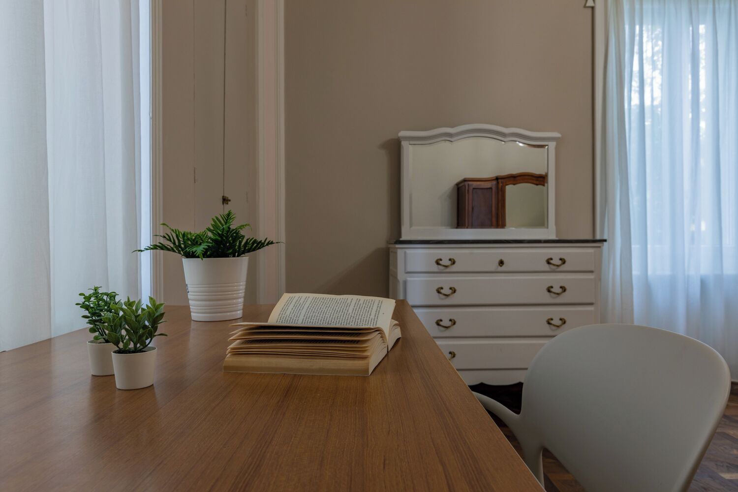 A room with a wooden table, a white chair, a dresser with a mirror, and two potted plants.