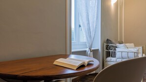 Una mesa de madera con un libro abierto, una silla, una ventana con cortinas traslúcidas y una cama con almohadas al fondo.