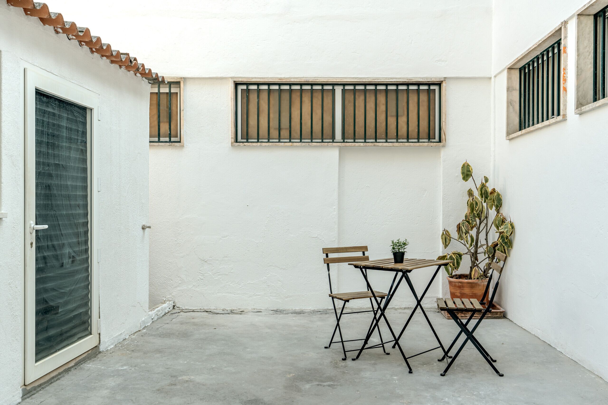 Une petite terrasse avec une table et des chaises, un pot de fleurs et une fenêtre grillagée.