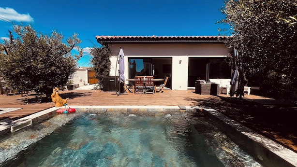 Une piscine avec une terrasse en bois, un espace patio aménagé avec du mobilier d’extérieur et un bâtiment doté d’un toit en tuiles.