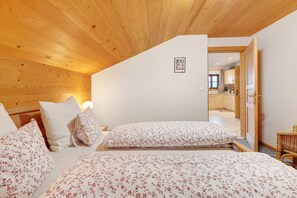 A bedroom with two beds, wooden ceiling, white walls, and a view into the kitchen.