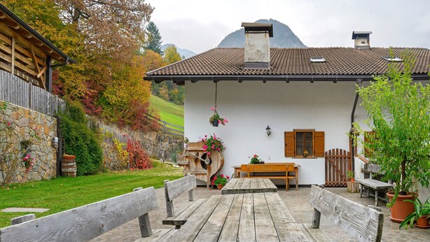 A rustic chalet with a stone wall, wooden benches, a wooden table, and hanging flower baskets.