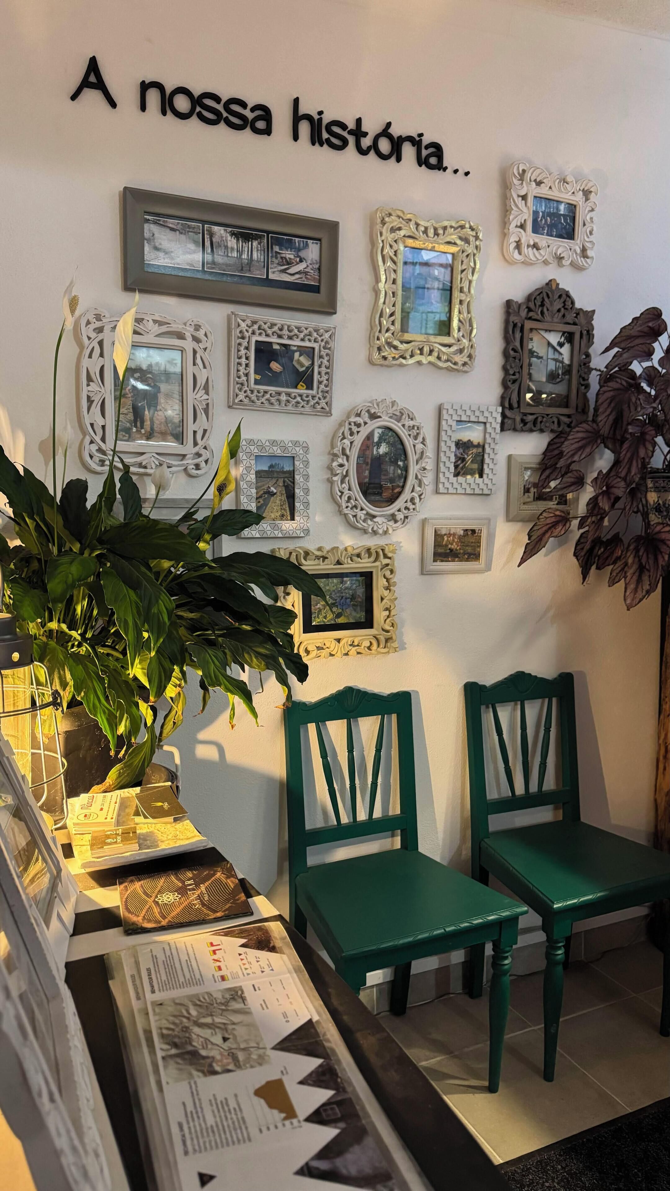 Een kamer met een muur versierd met verschillende ingelijste foto’s en een bordje met de tekst “A nossa história...”. Op de voorgrond staan twee groene stoelen en een potplant.