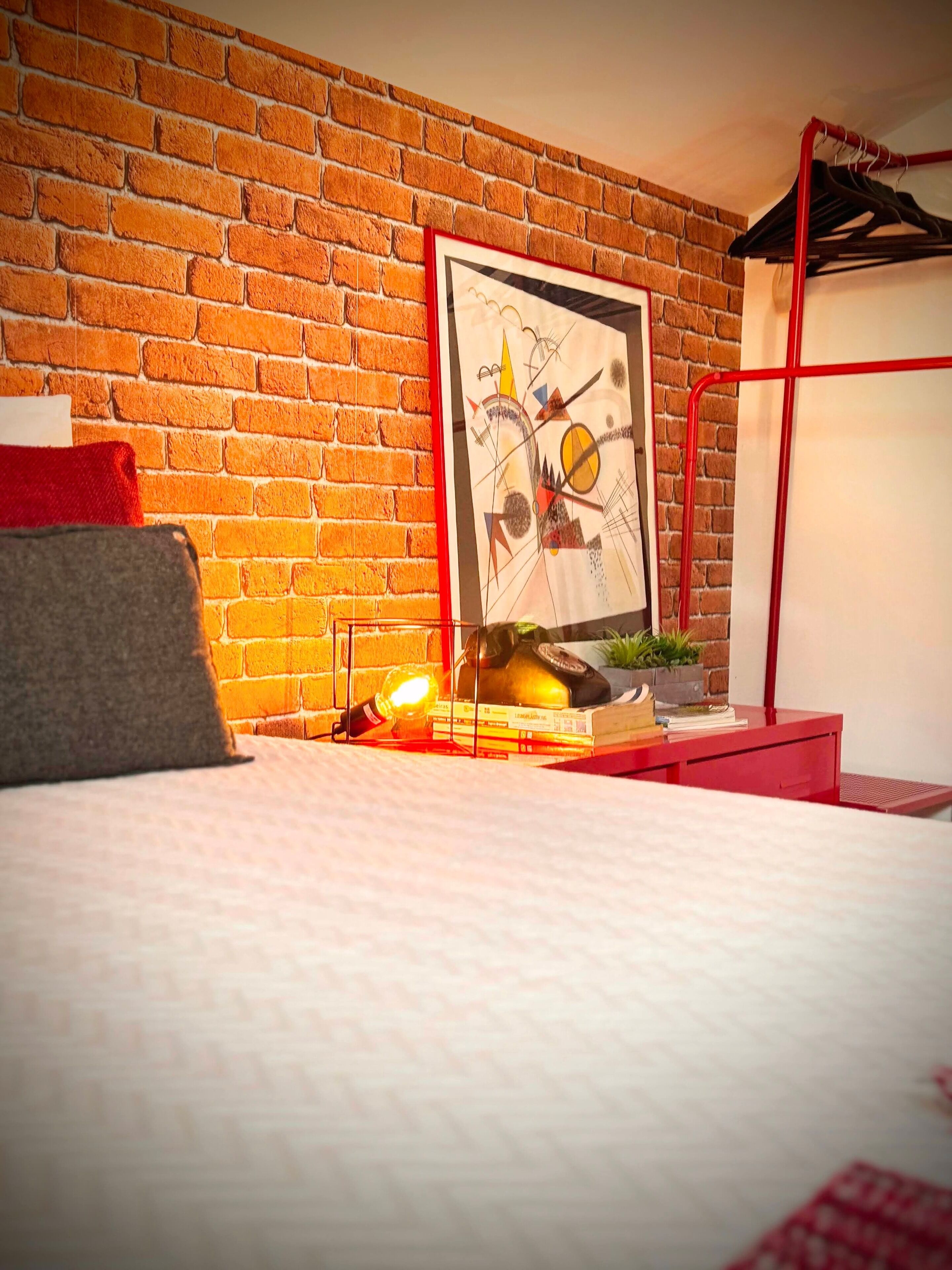 A room with a brick wall, a bed, a red desk, and a framed abstract painting.