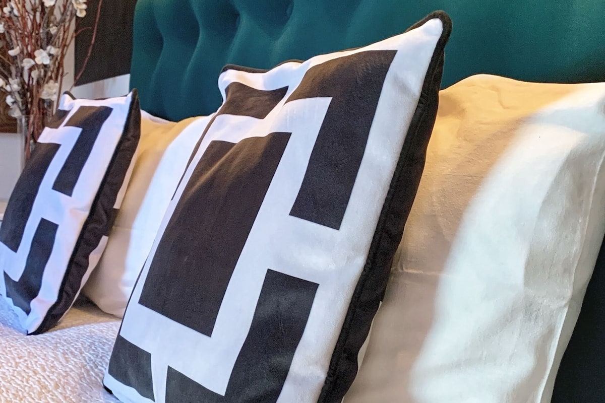 Una cama con cabecera acolchada, dos almohadas estampadas y una colcha con textura.