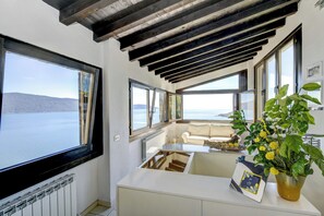 A room with a view of the sea, a white sofa, a table with a vase of flowers, and a framed picture.