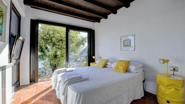 A bedroom with a bed, a window, a yellow lamp, and a view of greenery. - Lemon Garden Retreat - Villa Aqua (Gargnano)
