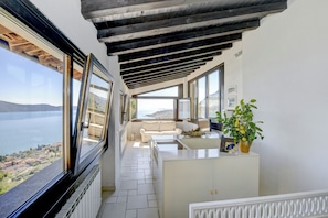 Een smal balkon met een witte bank, een klein tafeltje en een bloempot. Een groot raam biedt uitzicht op de zee en verre gebouwen.