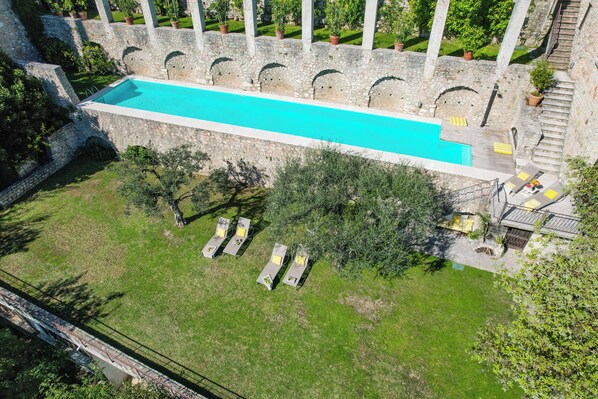 An aerial view of a swimming pool with a stone wall, surrounded by greenery and a few lounge chairs.