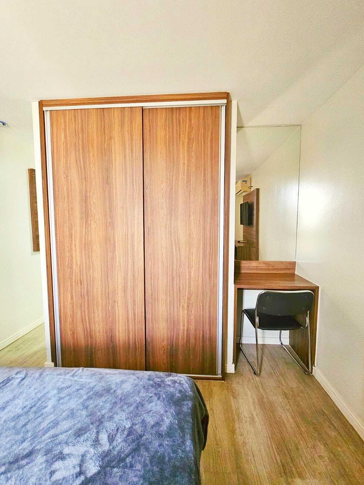 Une chambre avec une armoire en bois, un lit recouvert d’une couvre-lit à motifs, un bureau avec une chaise et un miroir accroché au mur.
