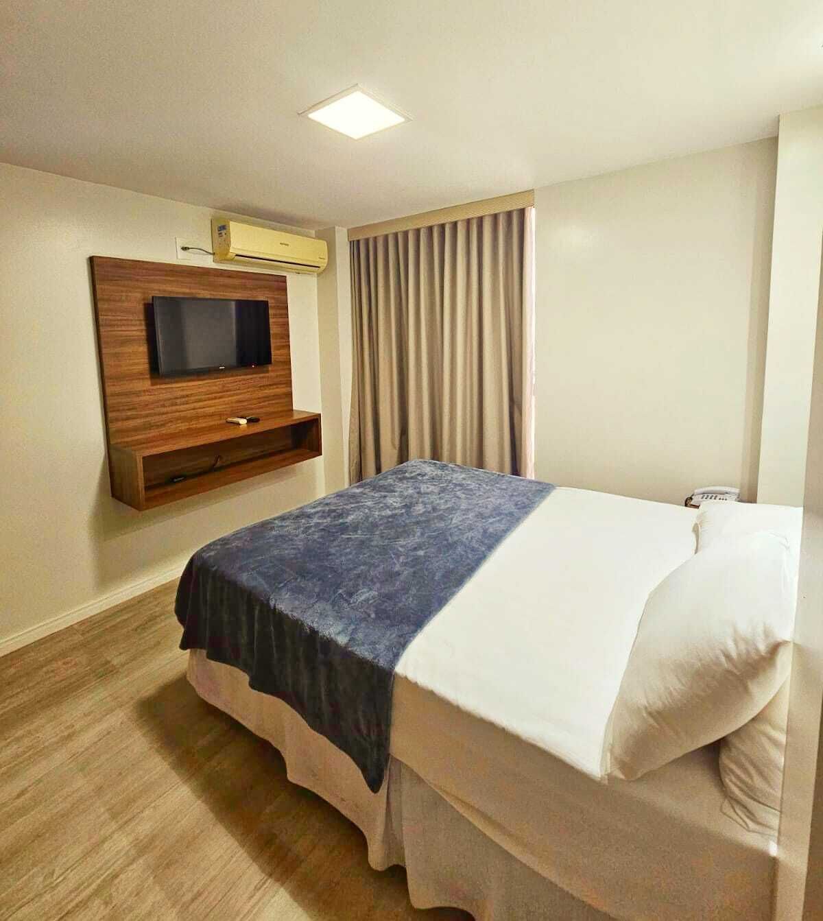 Una habitación de hotel con una cama, un televisor de pantalla plana montado en la pared, una ventana con cortinas y suelo de madera.