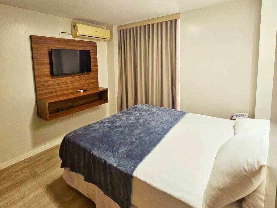 Una habitación de hotel con una cama, un televisor de pantalla plana montado en la pared, una ventana con cortinas y suelo de madera.