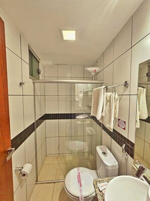 Ein modernes Badezimmer mit einer Duschkabine aus Glas, einer Toilette mit Hygienehinweis, einem Waschbecken mit Seifenspender und einem Handtuchhalter mit mehreren Handtüchern.
