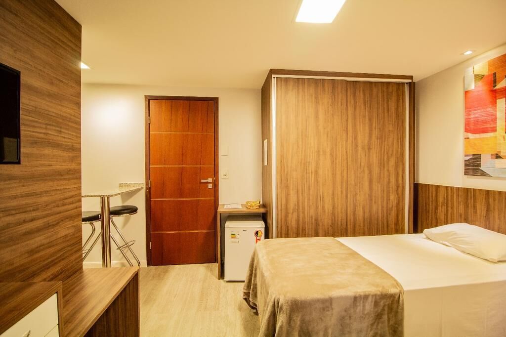 A hotel room with a bed, a desk, a chair, a wardrobe, and a small refrigerator.
