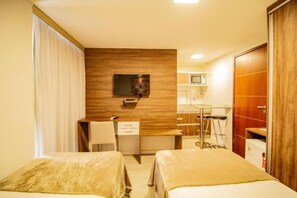 Ein Hotelzimmer mit zwei Betten, einem Schreibtisch, einem Stuhl, einem an der Wand montierten Fernseher und einer Küchenzeile.
