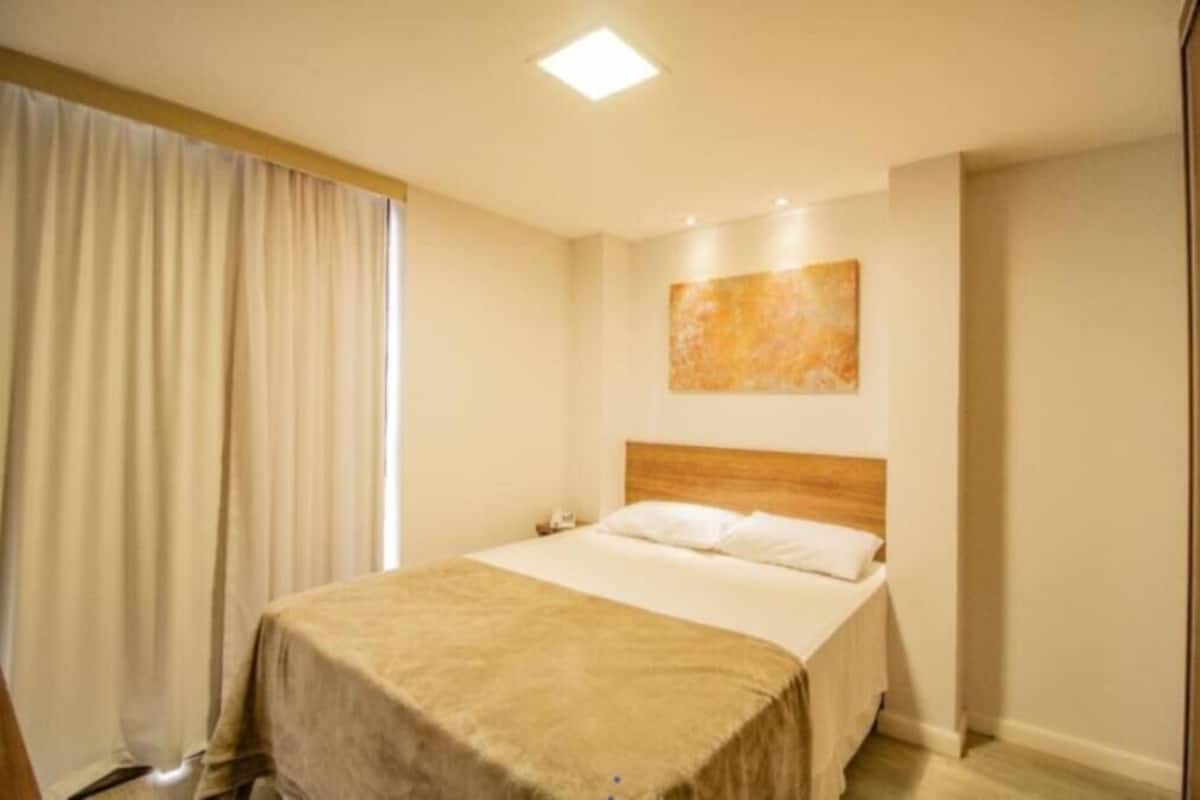 A hotel room with a bed, a nightstand, a painting on the wall, and a window with curtains.