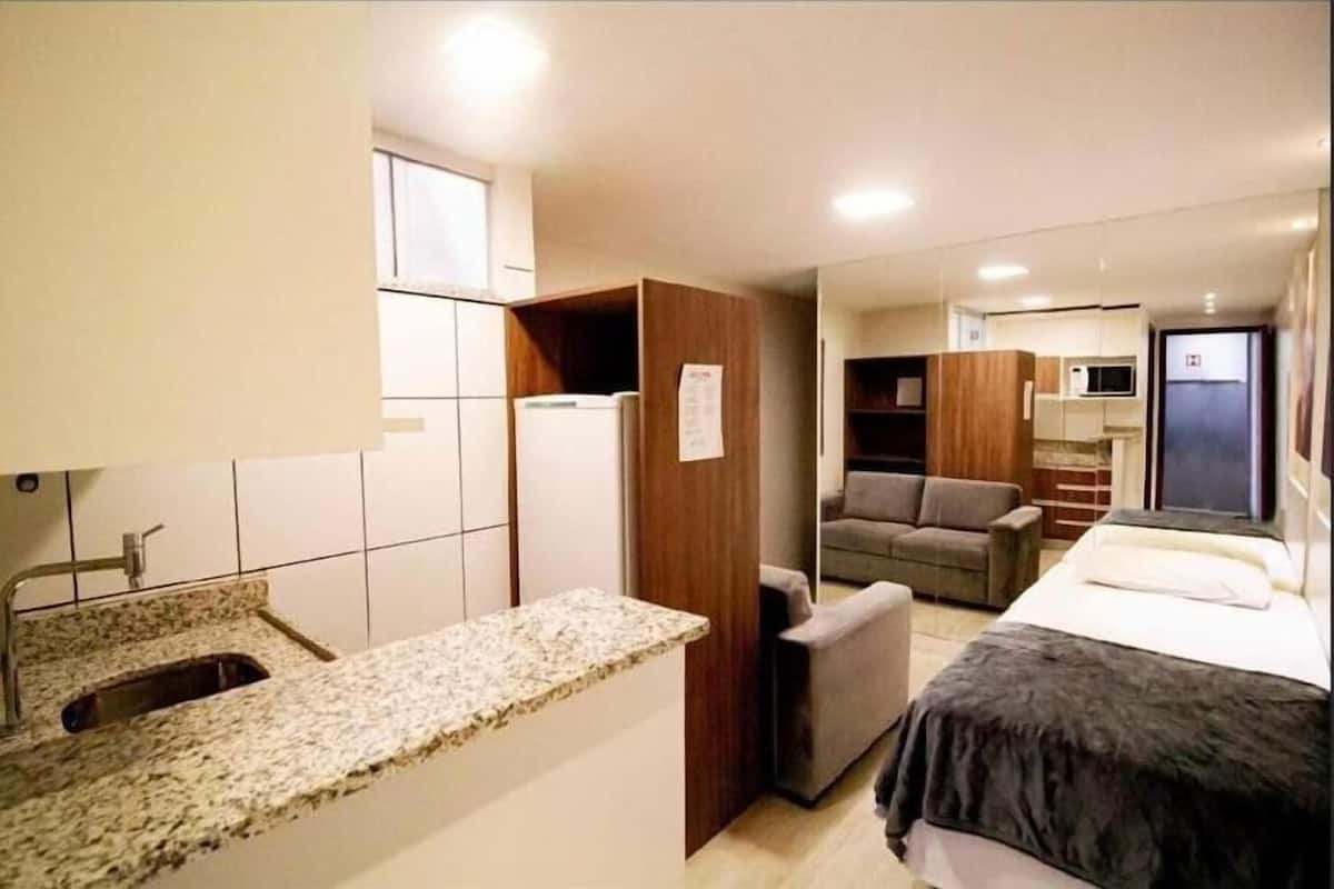 A compact hotel room with a kitchenette, a bed, a sofa, and a small seating area.