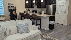 Un soggiorno moderno con un divano bianco, cuscini decorati, un tavolino rotondo e un angolo cottura con mobili scuri ed elettrodomestici in acciaio inossidabile.