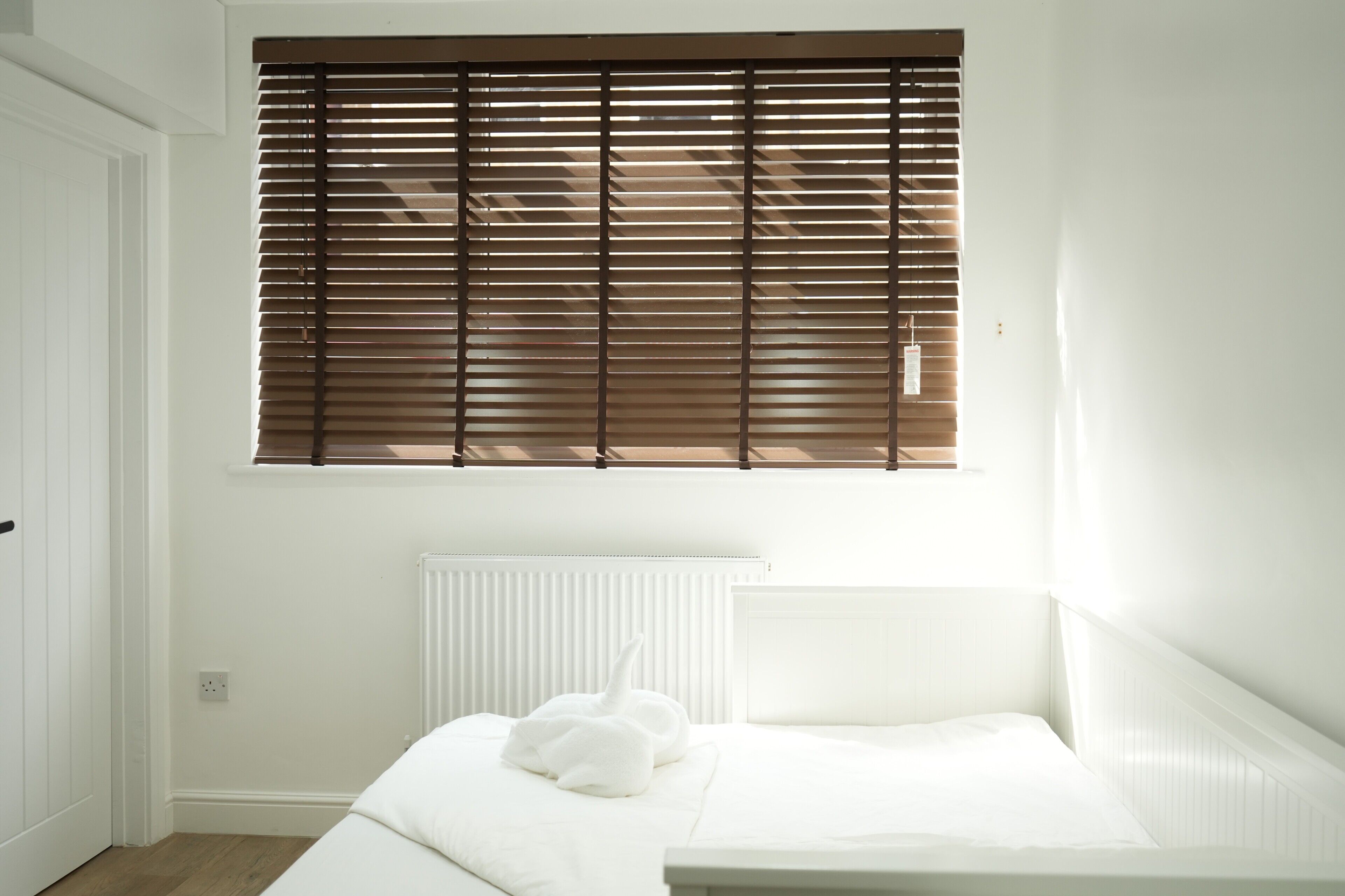 A room with a white bed, a white radiator, and a window with brown blinds.