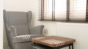 A room with a houndstooth patterned armchair, a wooden table, and a window with blinds. - Concept Rooms  (Enfield)