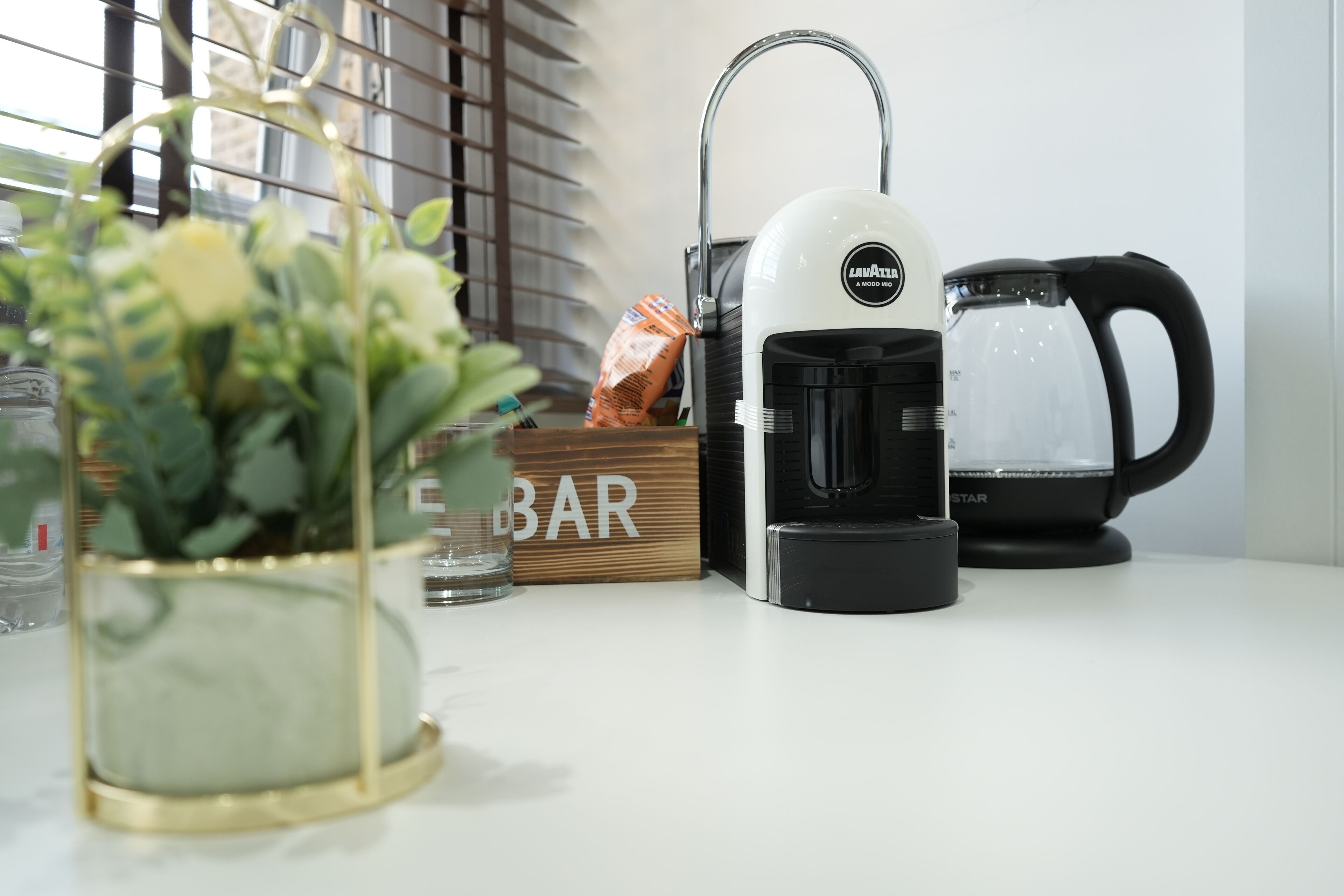 Una encimera de cocina con una cafetera, una jarra de vidrio, una planta en maceta, un caja de madera con la etiqueta 'BAR' y una ventana con persianas.