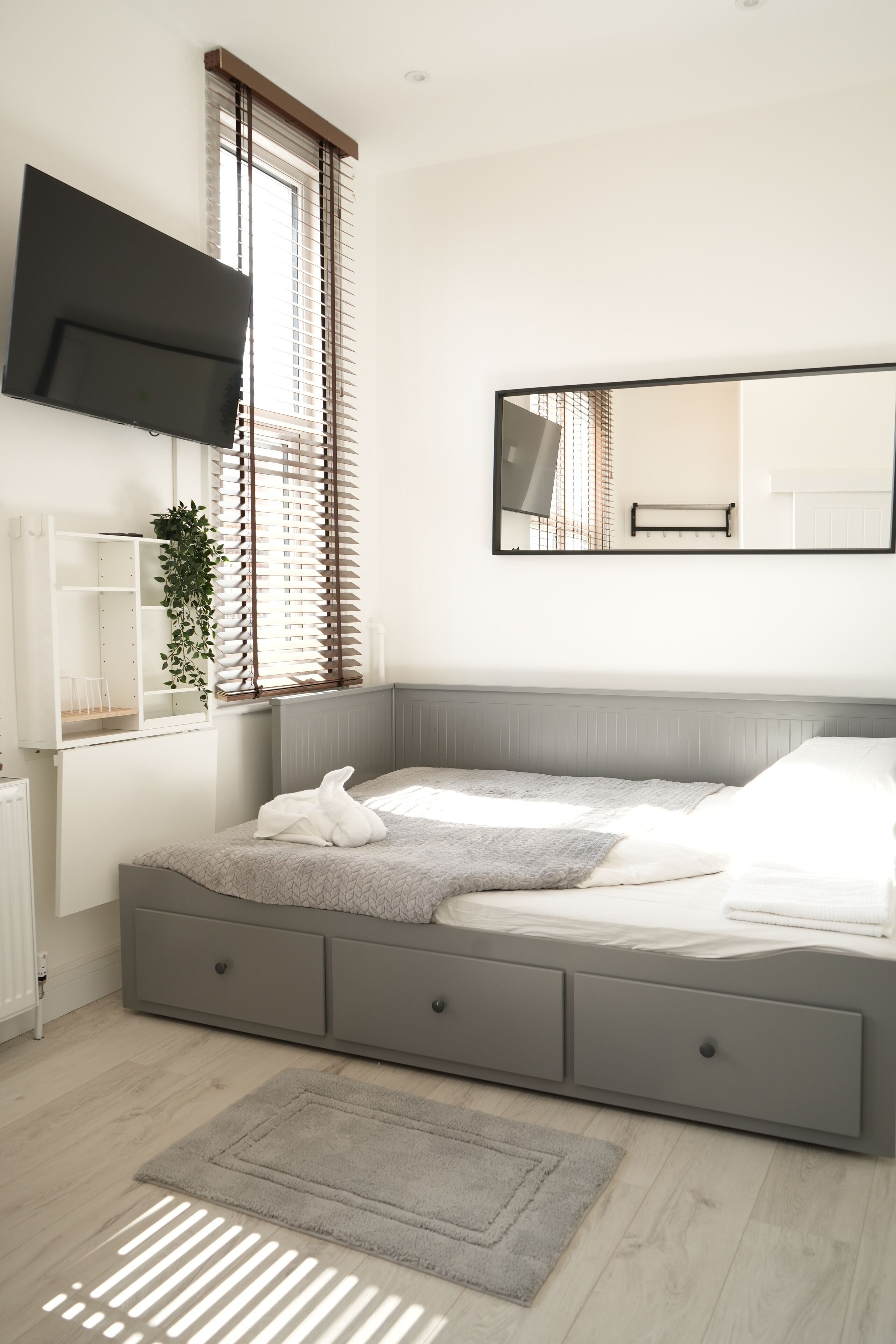 Ein modernes Schlafzimmer mit einem großen Bett, einem an der Wand montierten Fernseher und einem Fenster mit Jalousien.