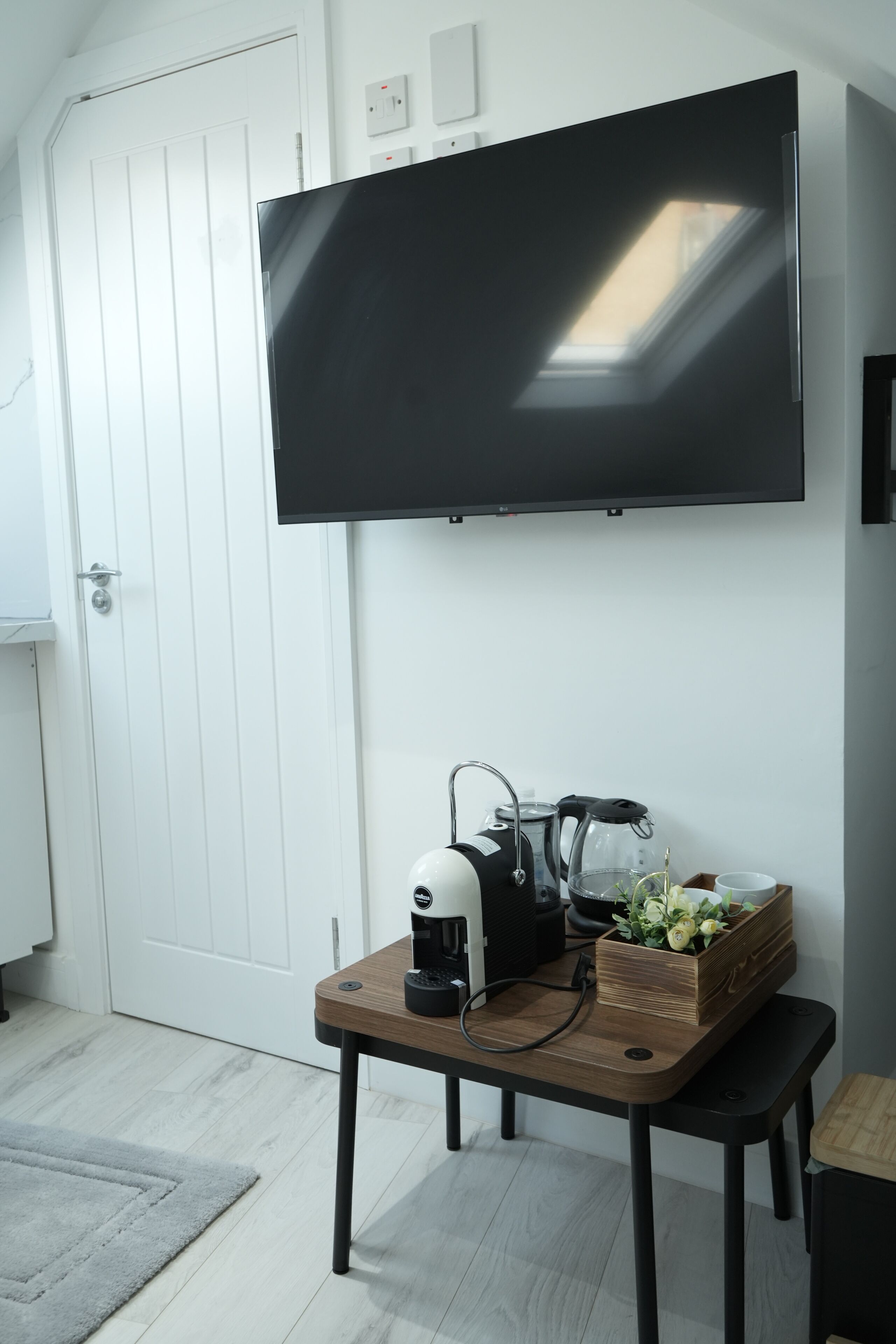 A modern living room with a flat-screen TV mounted on the wall, a coffee maker on a wooden table, and a small bench.