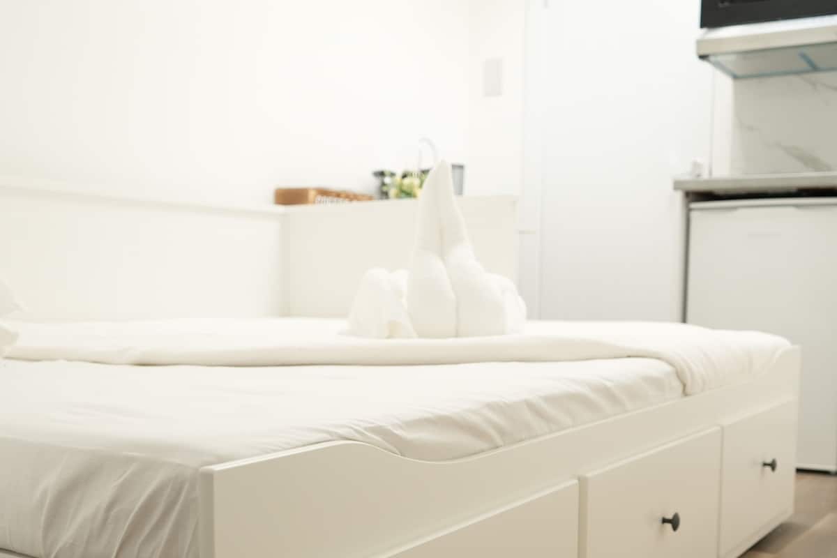 A white bedroom with a bed, a nightstand, and a microwave above a white cabinet.