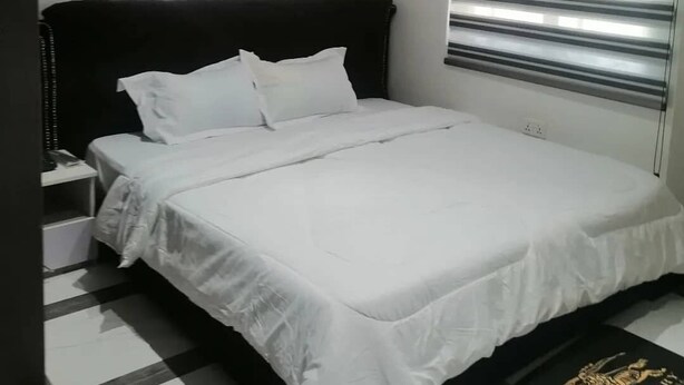 Un dormitorio con cabecera negra, ropa de cama blanca y dos ventanas con persianas horizontales.