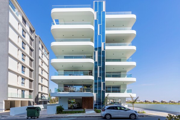 A modern multi-story building with balconies, a car parked in front, and a green trash bin. - Aelia Chic 2-BR Apt & Balcony in Larnaca (Larnaca)