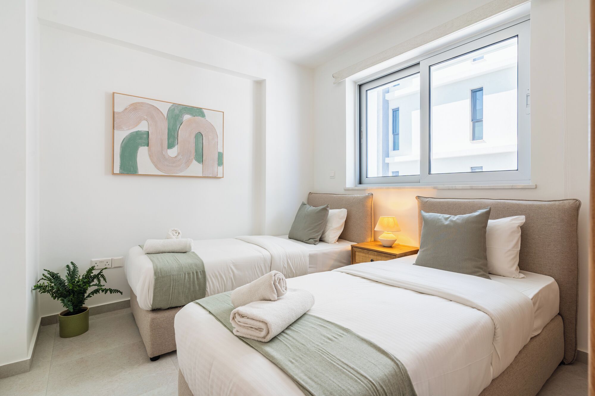 Hotellihuone, jossa on kaksi sänkyä, ikkuna, josta avautuu näkymä rakennuksiin, yöpöytä lampulla ja seinälle kiinnitetty abstrakti taulu.
