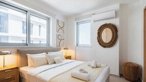 Un dormitorio moderno con una cama grande, mesitas de noche, un espejo redondo y vistas de la ciudad a través de amplias ventanas.