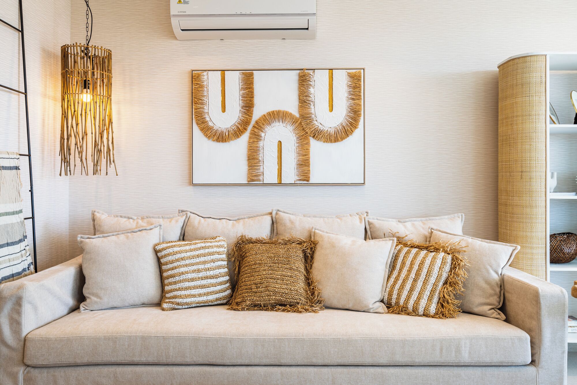 A neutral-toned living room with a beige sofa, several cushions, a hanging light fixture, and a framed abstract artwork on the wall.